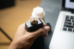 Hand holding a vape mod with exposed heated coil near a laptop