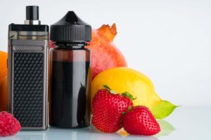 Nicotine salt vape device and e-liquid bottle surrounded by fresh fruit including pomegranate, lemon, and strawberries